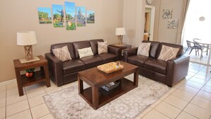 A living room with a brown leather sofa, two side tables with lamps, a coffee table, and a rug. There are wall-mounted pictures and a clock on the wall. - Family friendly, 10 mins to Disney, WIFI, Resort style amenities, Backyard pool (Kissimmee)