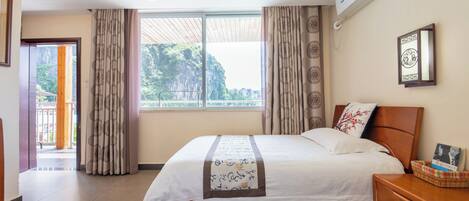 Una habitación de hotel con una cama grande, una mesita de noche, una ventana con cortinas, un aire acondicionado de pared y un cuadro enmarcado.