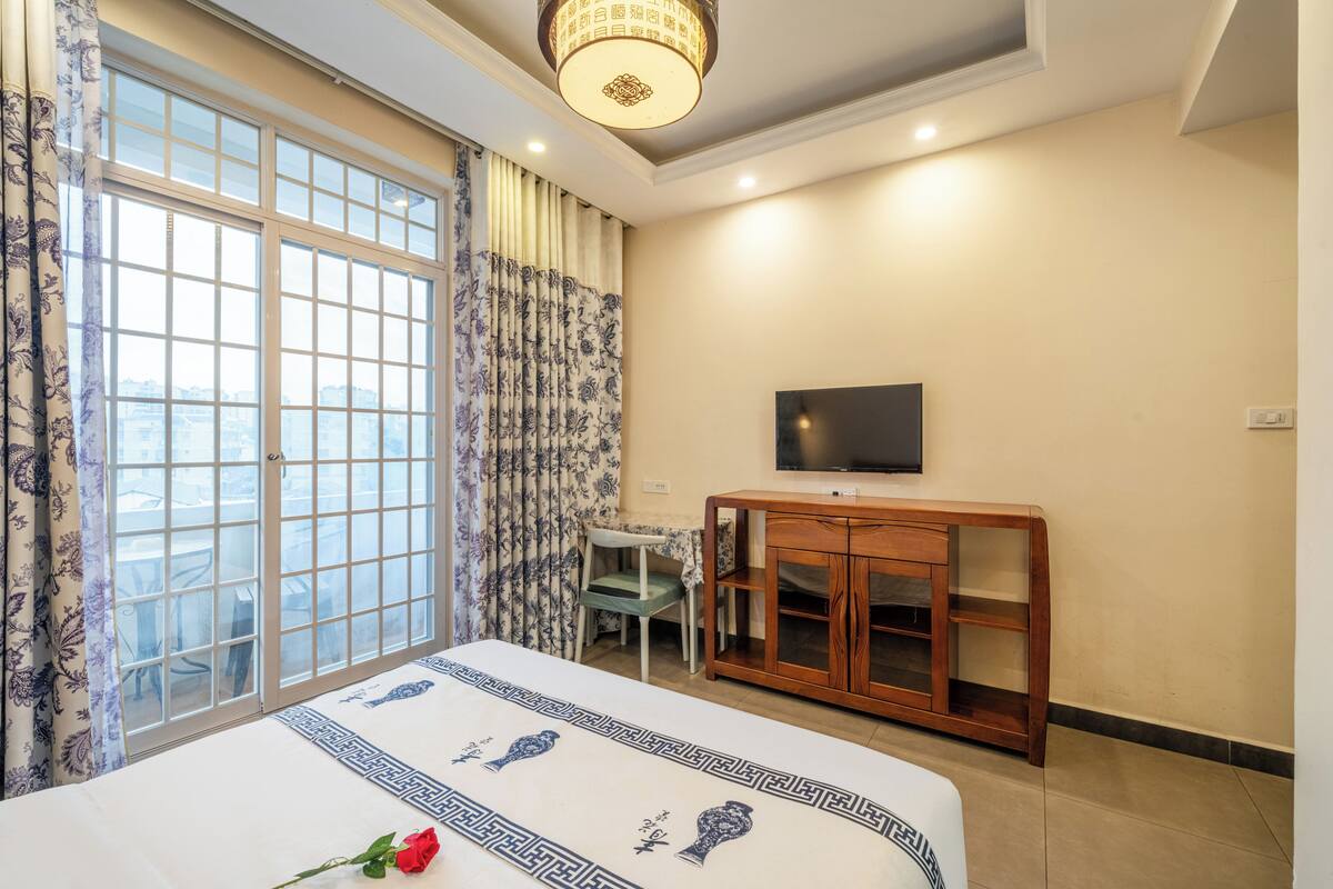 A hotel room with a large window, a bed with a floral bedspread, a wooden cabinet, a television on the wall, and a small table with a chair.