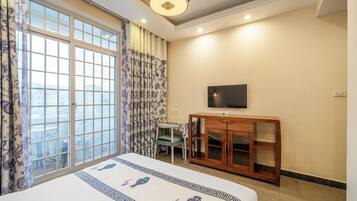 Habitación de hotel con un ventanal grande, una cama con cubrecama floral, un armario de madera, un televisor en la pared y una pequeña mesa con una silla.