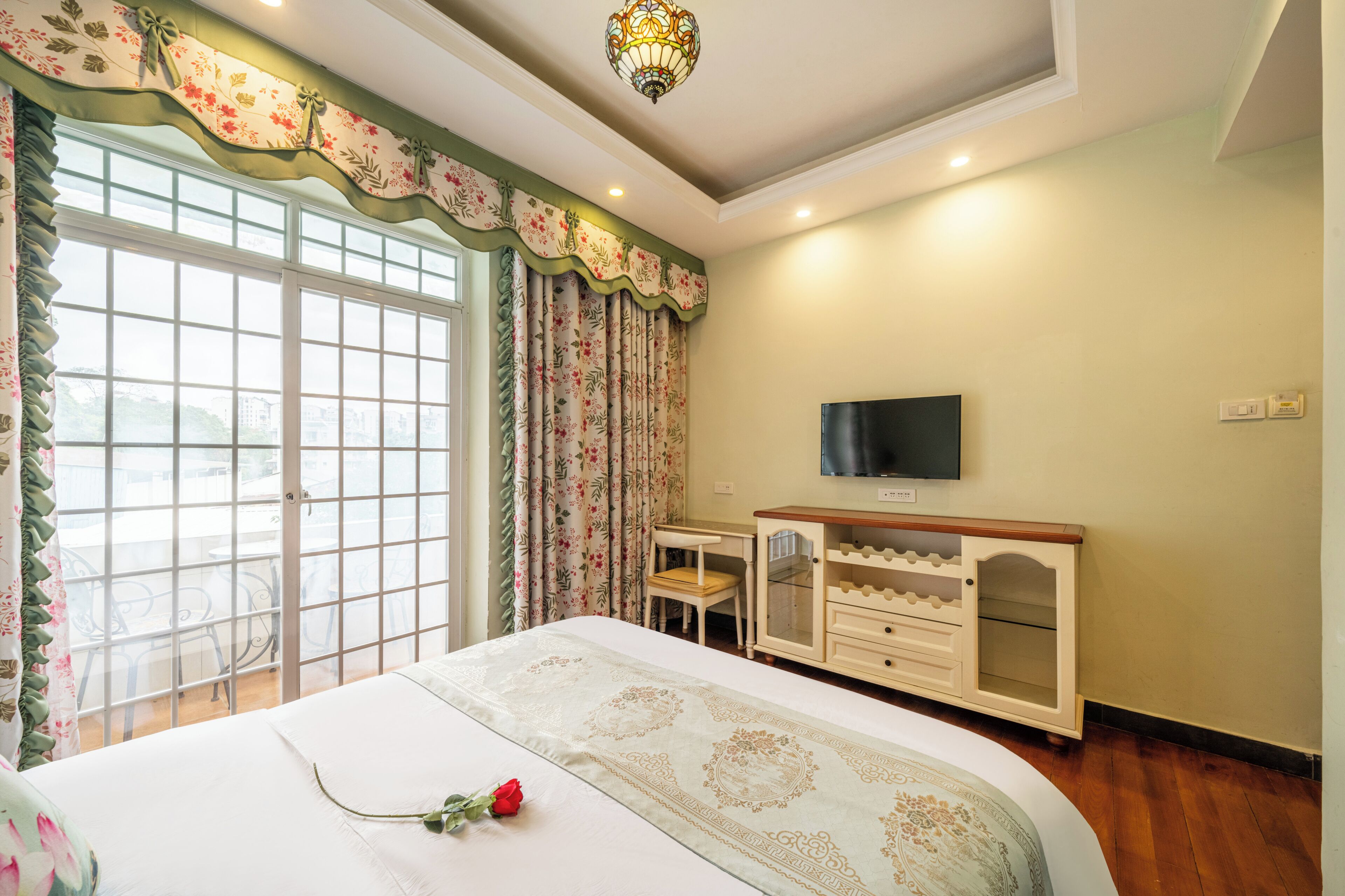 A bedroom with a large window, a bed with a decorative bedspread, a desk with a chair, a television, and a cabinet.