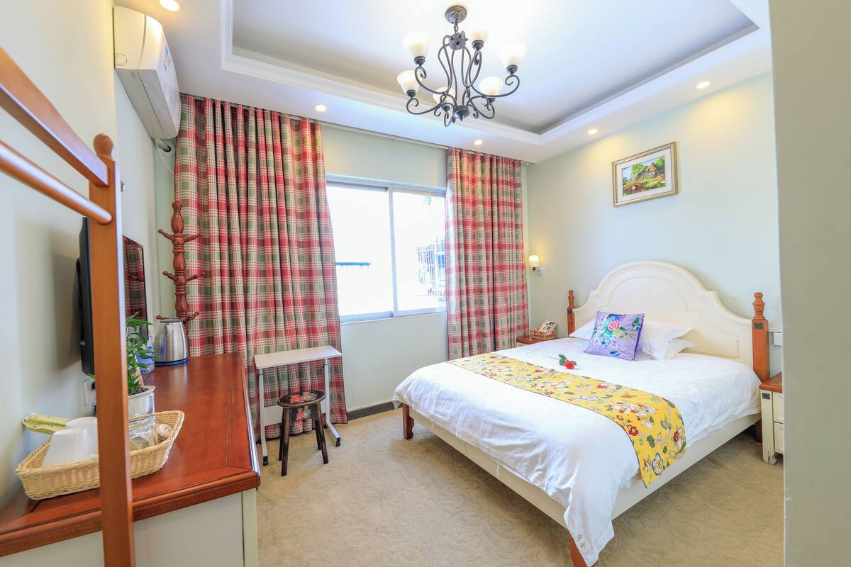 A bedroom with a bed, a desk, a chair, a window with curtains, a chandelier, and a picture on the wall.
