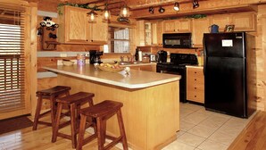 A kitchen with wooden cabinets, a black refrigerator, a microwave, a stove, and a sink. There are three wooden stools and a basket of fruit on the counter. - dream catcher. Beautiful Mountain Veiws and Real Log Cabin Charm! (Sevierville)