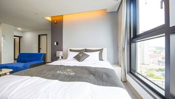 Habitación de hotel con una cama, una silla azul, una mesita de noche, una ventana con vista a la ciudad y una lámpara de pared.