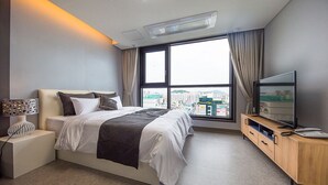 A modern hotel room with a large bed, a TV, a desk, and a view of the city through large windows.