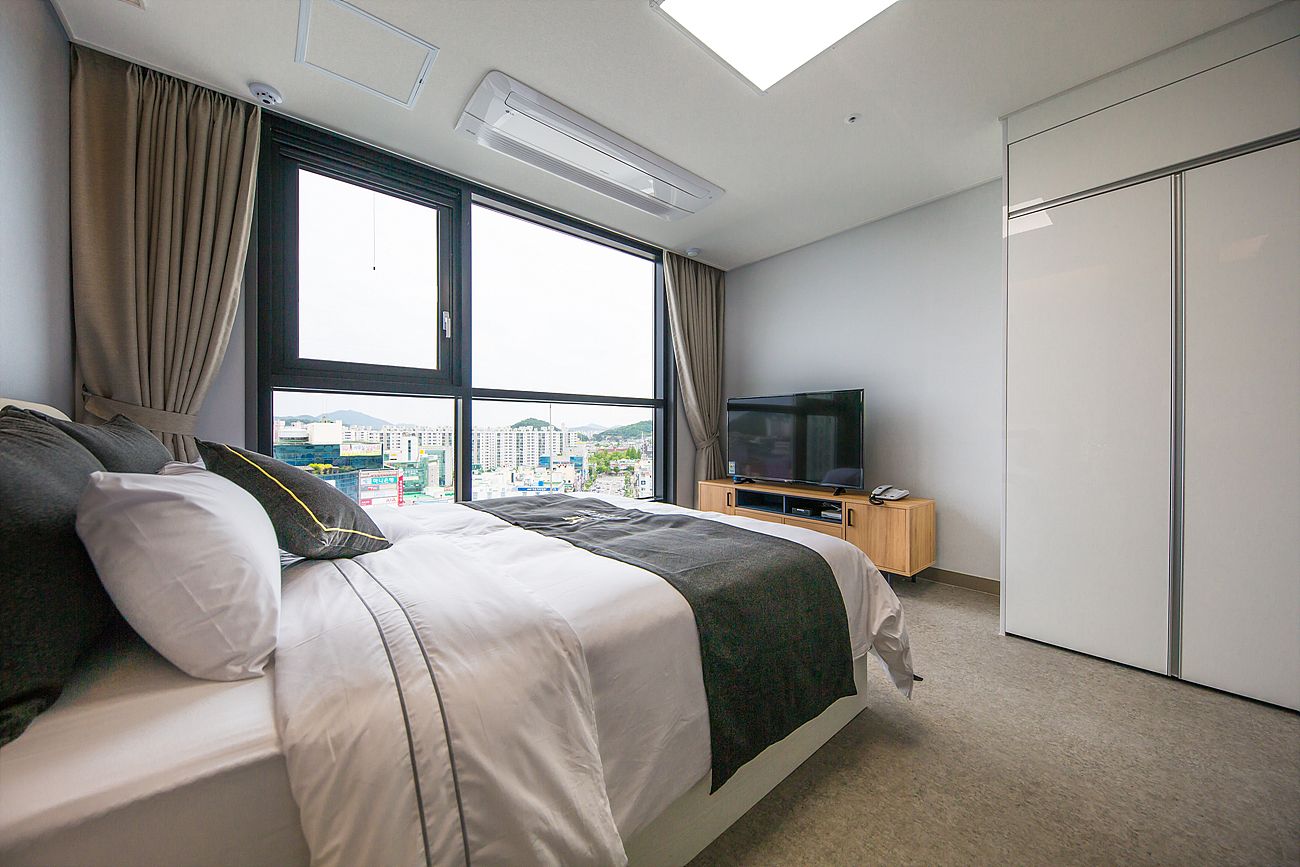 Quarto de hotel com uma cama grande, televisão, guarda-roupa e vista para a cidade pela janela.