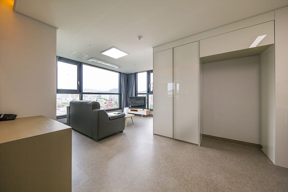 A modern hotel room with a large window, a grey sofa, a flat-screen TV, and a wardrobe.