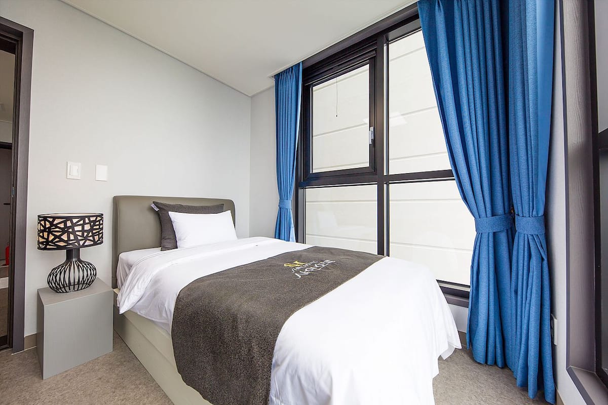A neatly made bed with a dark blanket, a bedside lamp, and a window with blue curtains.