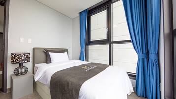 A neatly made bed with a dark blanket, a bedside lamp, and a window with blue curtains.