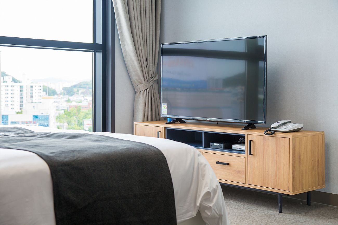 Quarto de hotel com cama, televisão sobre um móvel de madeira e vista para a cidade pela janela.