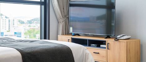 A hotel room with a bed, a TV on a wooden cabinet, and a city view through the window.