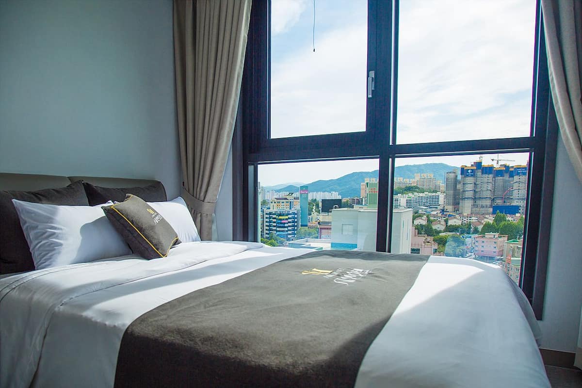 A hotel room with a large bed, a view of a cityscape through a window, and a curtain on the left side.