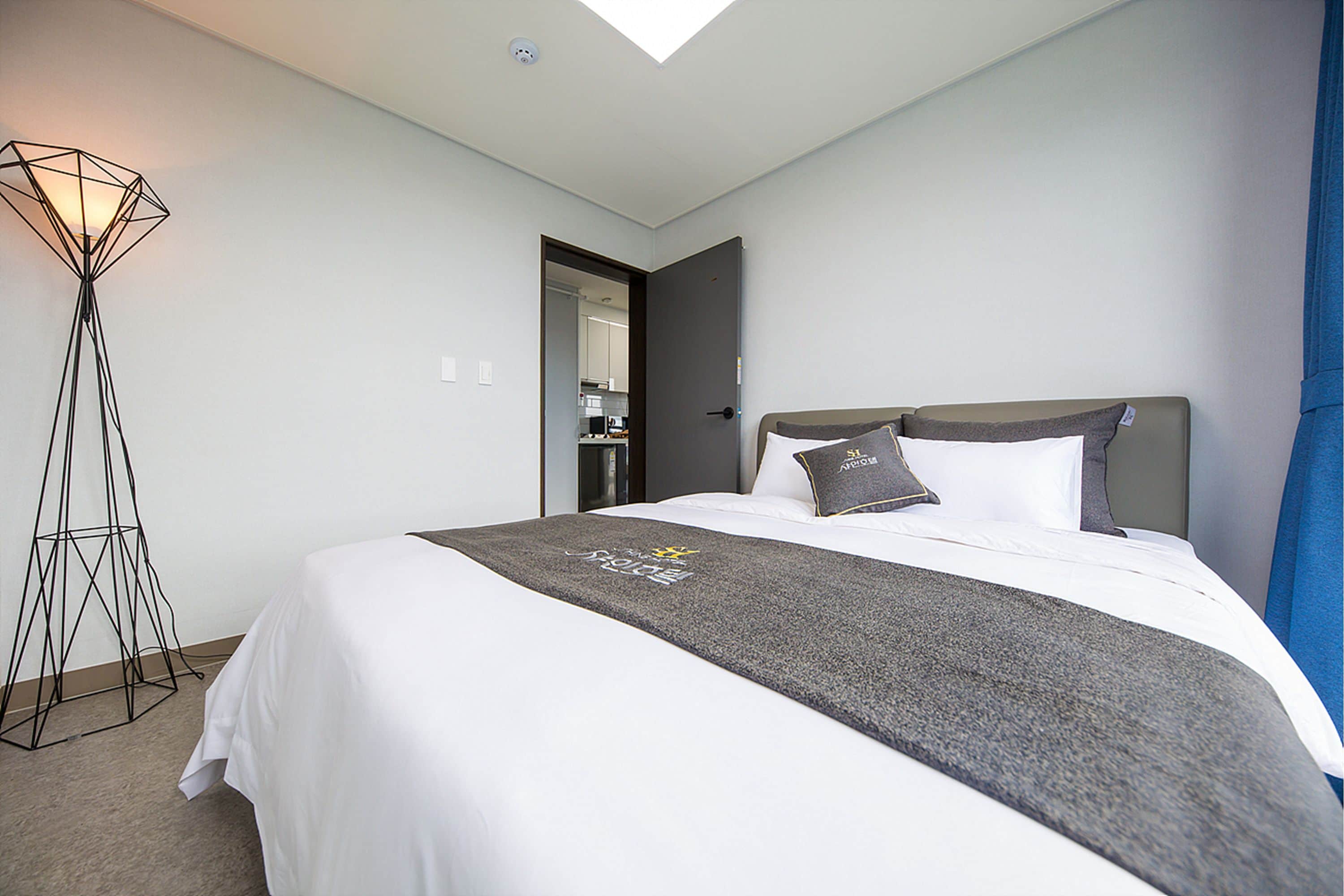 A hotel room with a neatly made bed, a floor lamp, and a door leading to a kitchen area.