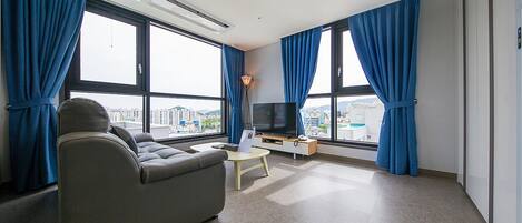 A modern hotel room with a sofa, a TV, and large windows with curtains.