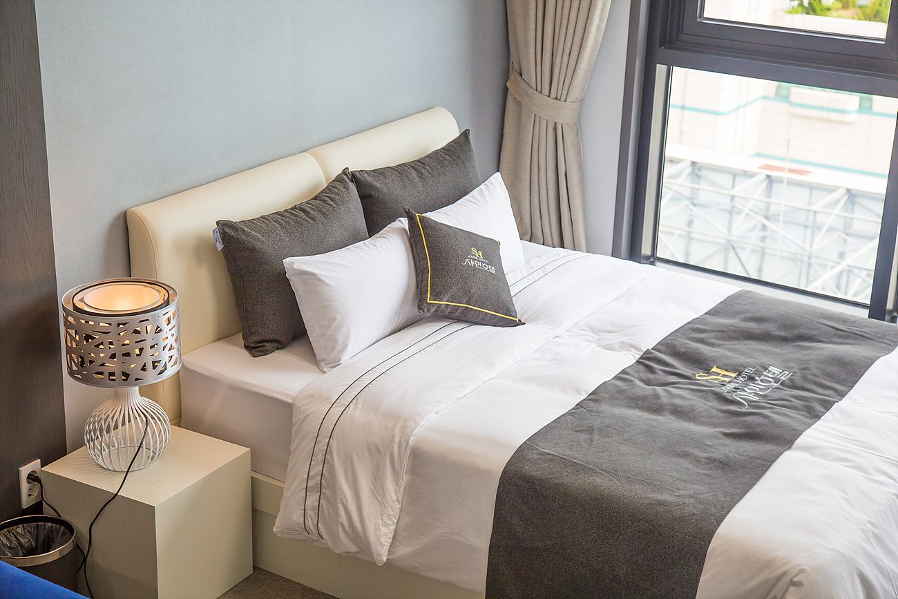 Una cama bien hecha con una colcha gris y blanca, dos almohadas y una lámpara de noche sobre una mesita blanca.