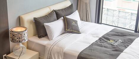 Una cama bien hecha con una colcha gris y blanca, dos almohadas y una lámpara de noche sobre una mesita blanca.