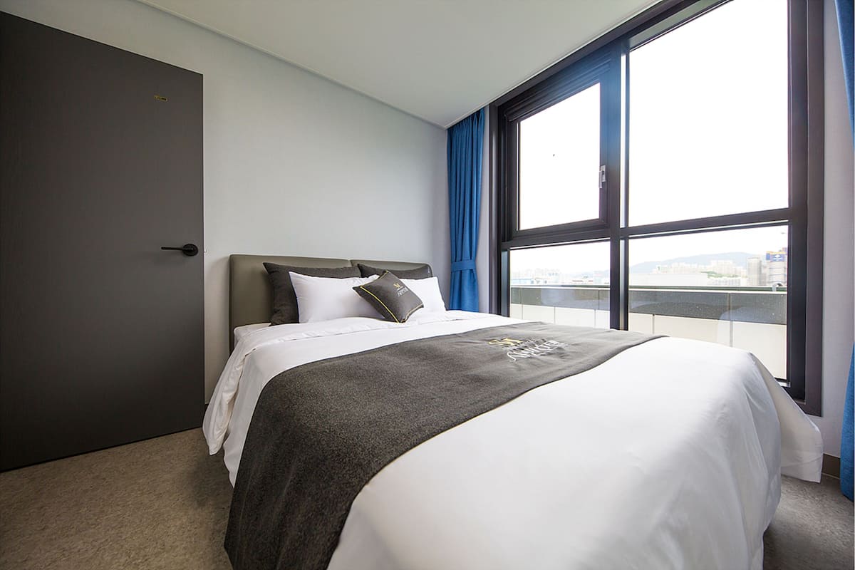 A hotel room with a large bed, a view of the city through the window, and a door to the left.