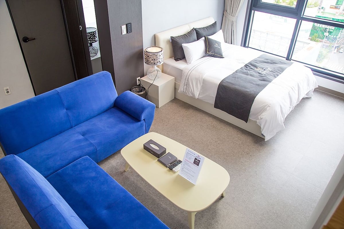 A hotel room with a blue sofa, a small table with a lamp, a bed with white bedding, and a view through the window.