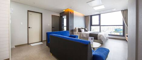 A modern hotel room with a sofa, a bed, a small table, and a large window offering a city view.