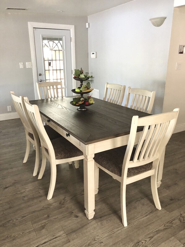 Un comedor con una mesa de madera y seis sillas, un centro de mesa decorativo con frutas y una puerta con ventana.