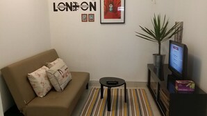 A living room with a beige sofa, a small black side table, a striped rug, a potted plant, a TV on a stand, and a framed 'LONDON' sign on the wall.