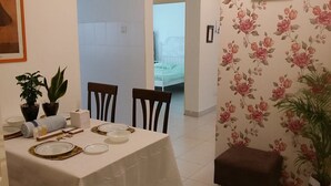 A dining area with a table set for two, two chairs, a small potted plant, and a brown ottoman.