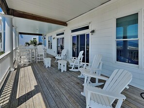 A covered outdoor deck with white Adirondack chairs and a small table, wooden flooring, and large windows. - Oceans 111 | Oceanfront with Elevator + Direct Beach Access! (Topsail Beach)