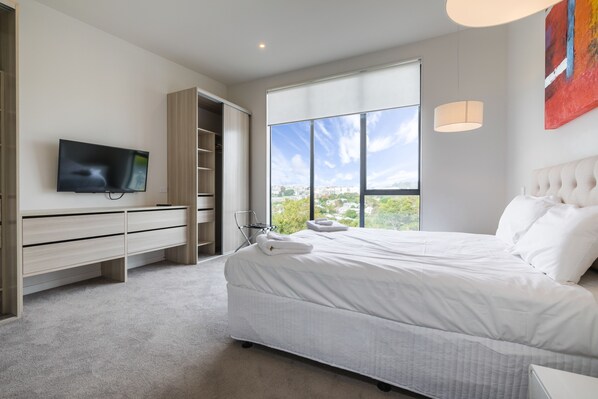 A modern bedroom with a large bed, a TV, a wardrobe, and a window with a city view.