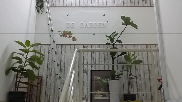 Una escalera con barandilla que conduce a una pared de madera con 'DE GARDEN' grabado. Hay plantas en macetas en los escalones y un cuadro enmarcado en la pared.