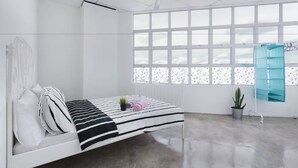 Una habitación luminosa y blanca con una cama, un ventilador de techo, un ventanal grande y una planta en maceta.