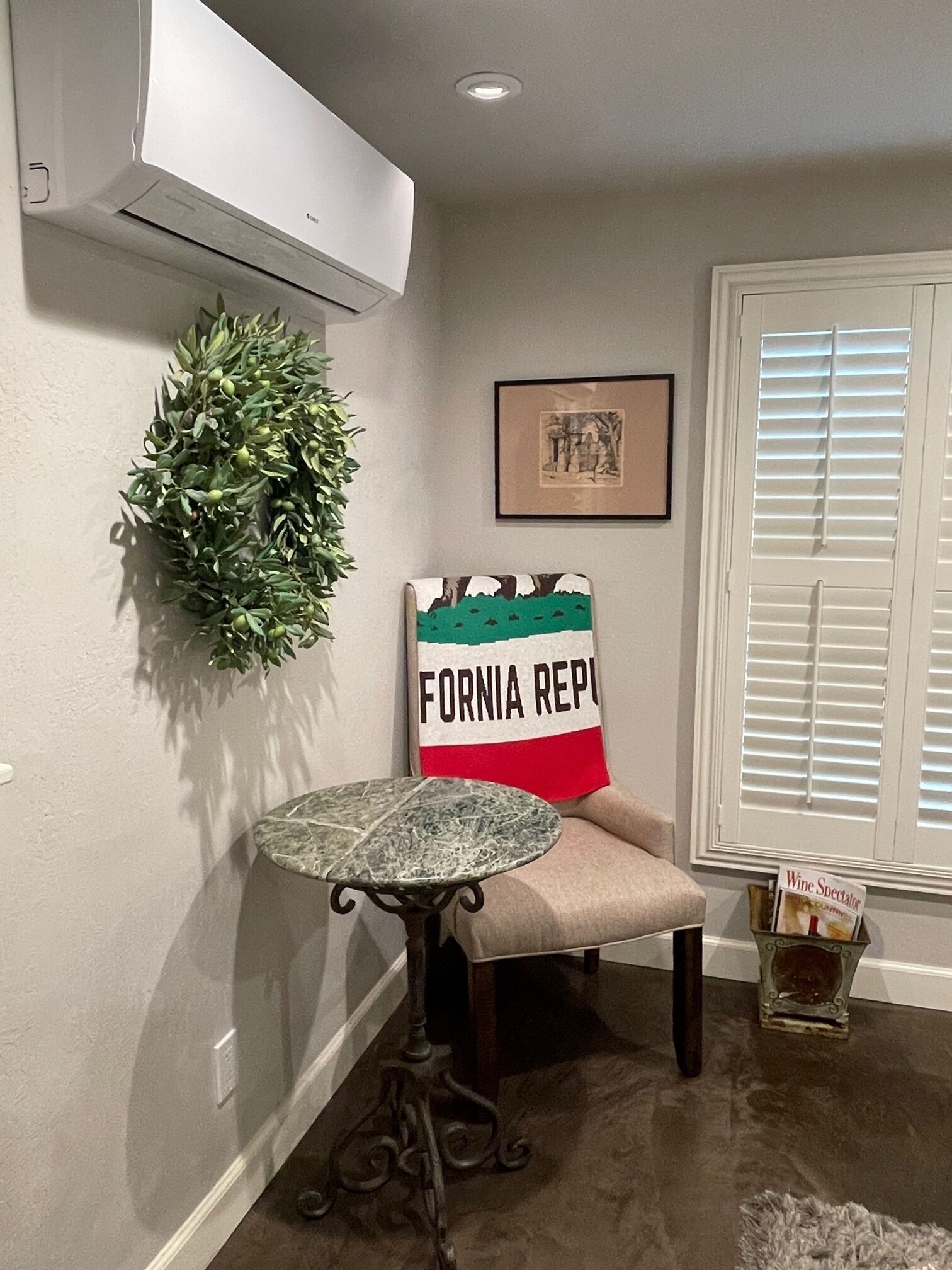 A corner room with a wall-mounted air conditioner, a potted plant, a framed picture, a chair with a pillow, a small table, and a window with blinds.