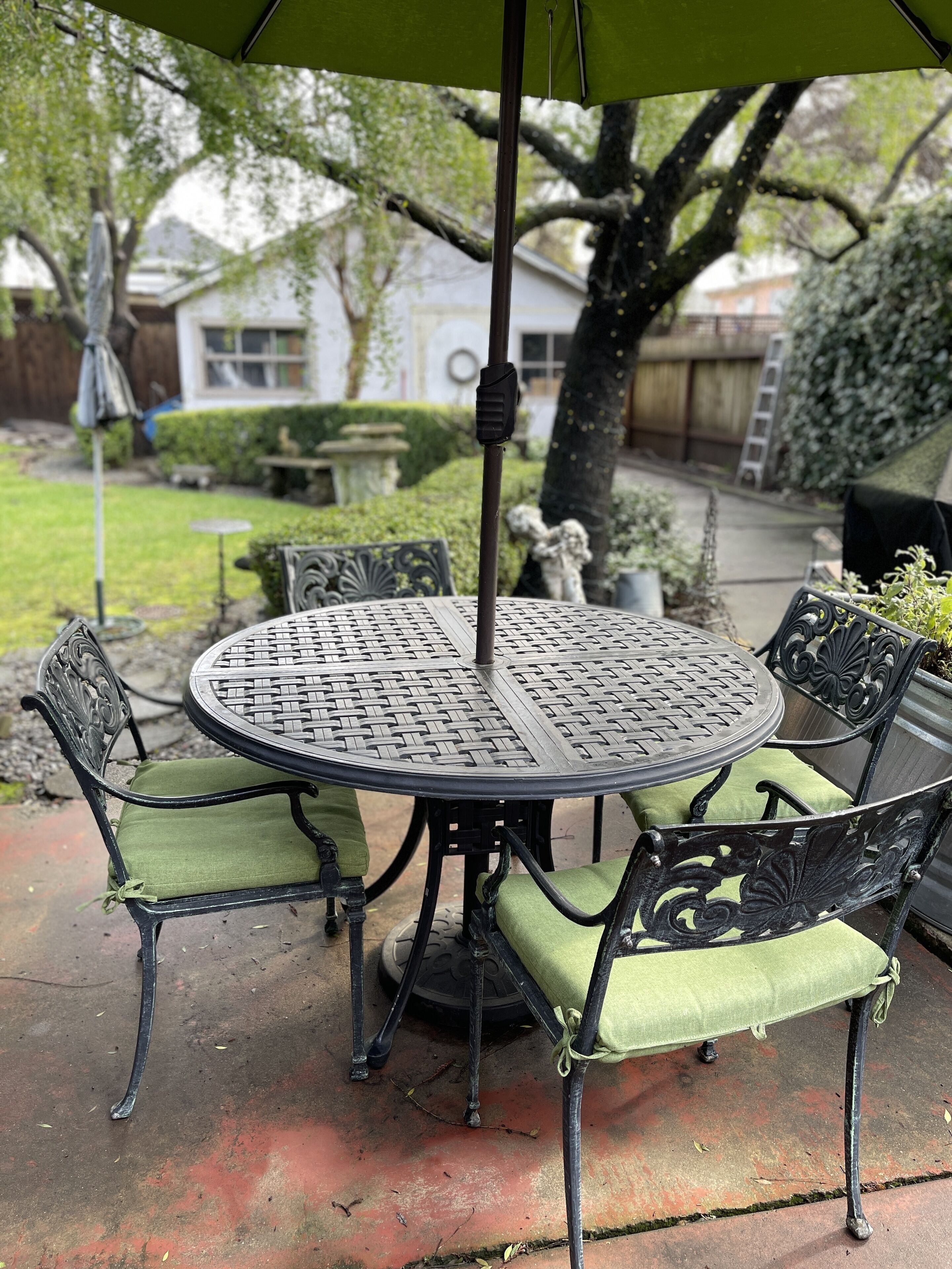 A round metal table with green cushioned chairs, an umbrella, and a decorative metal pole.