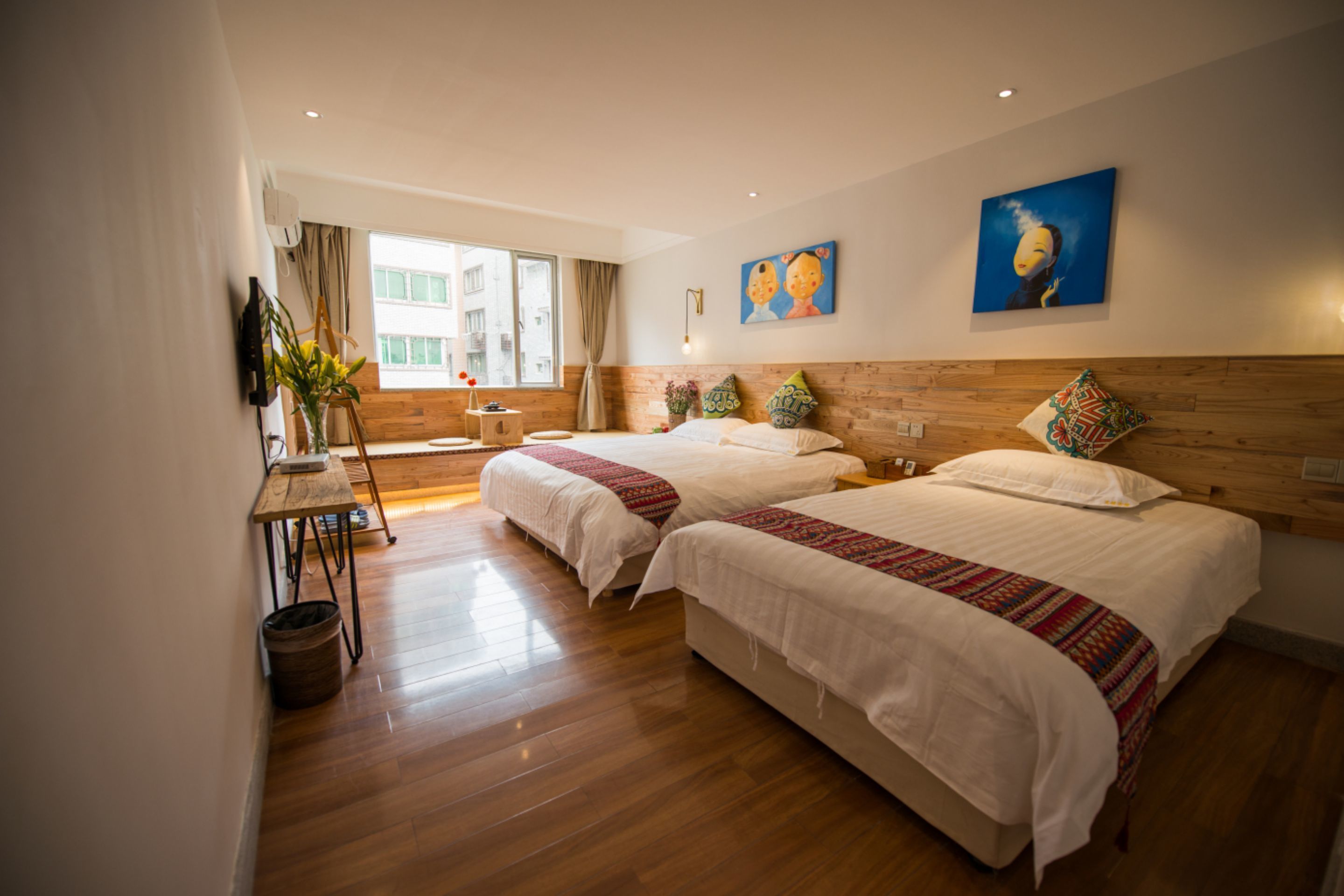 A hotel room with two beds, wooden flooring, a desk, and a window with curtains.