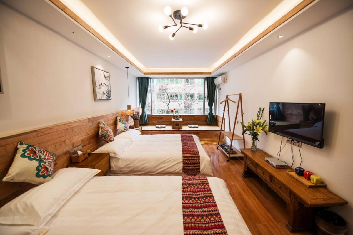 A hotel room with two beds, a TV, a wooden bench, and a window with curtains.