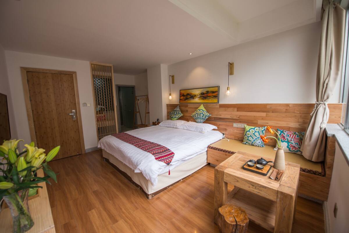 A bedroom with a wooden bed, bedside table with tea set, and a window with curtains.