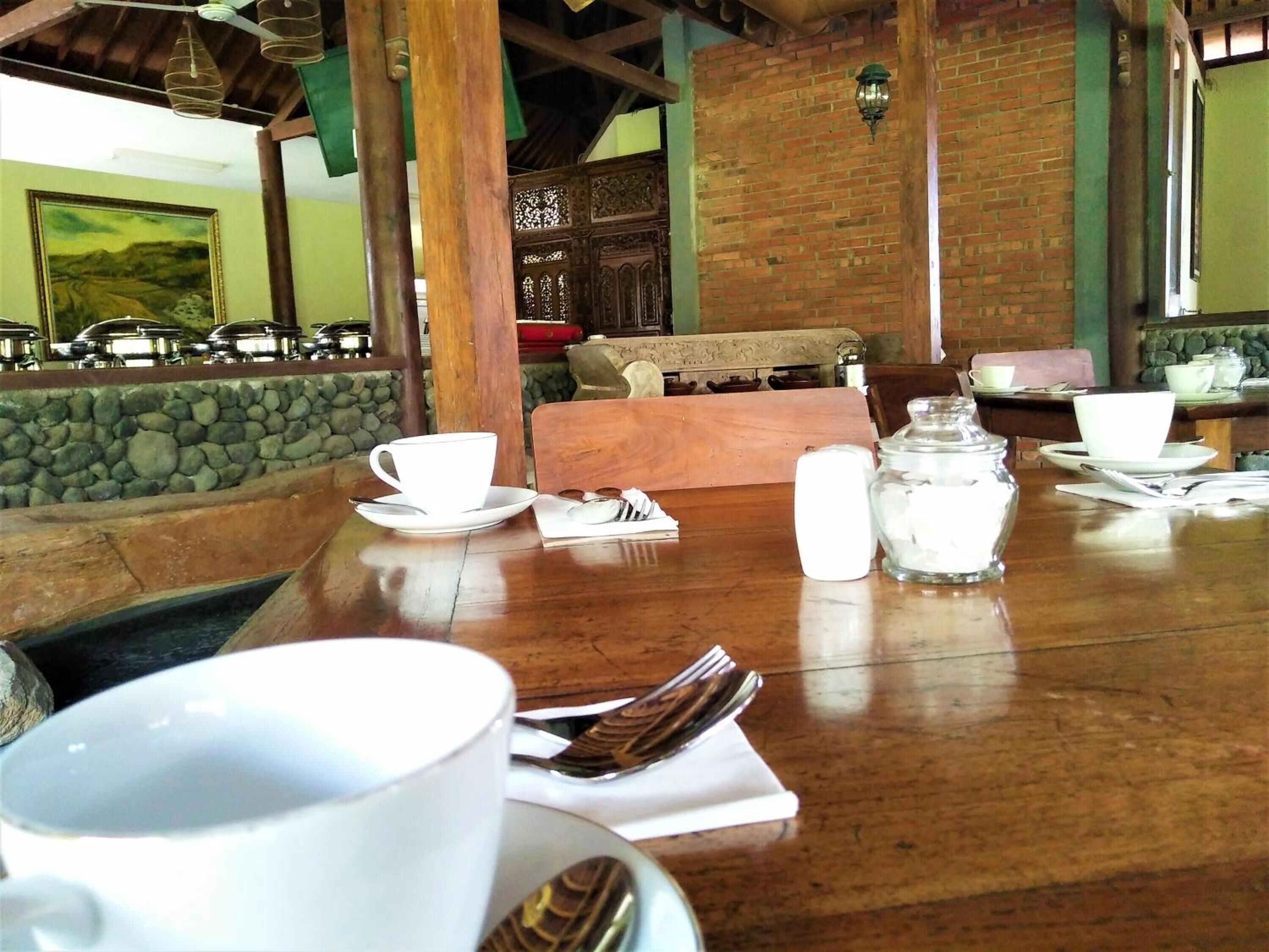 A dining table set with white cups, saucers, and cutlery, with a stone wall and wooden furniture in the background.