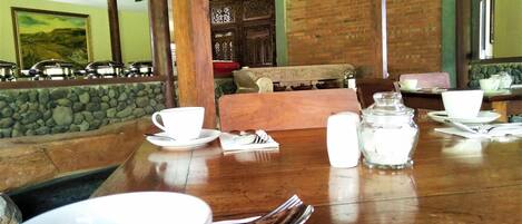 A dining table set with white cups, saucers, and cutlery, with a stone wall and wooden furniture in the background.