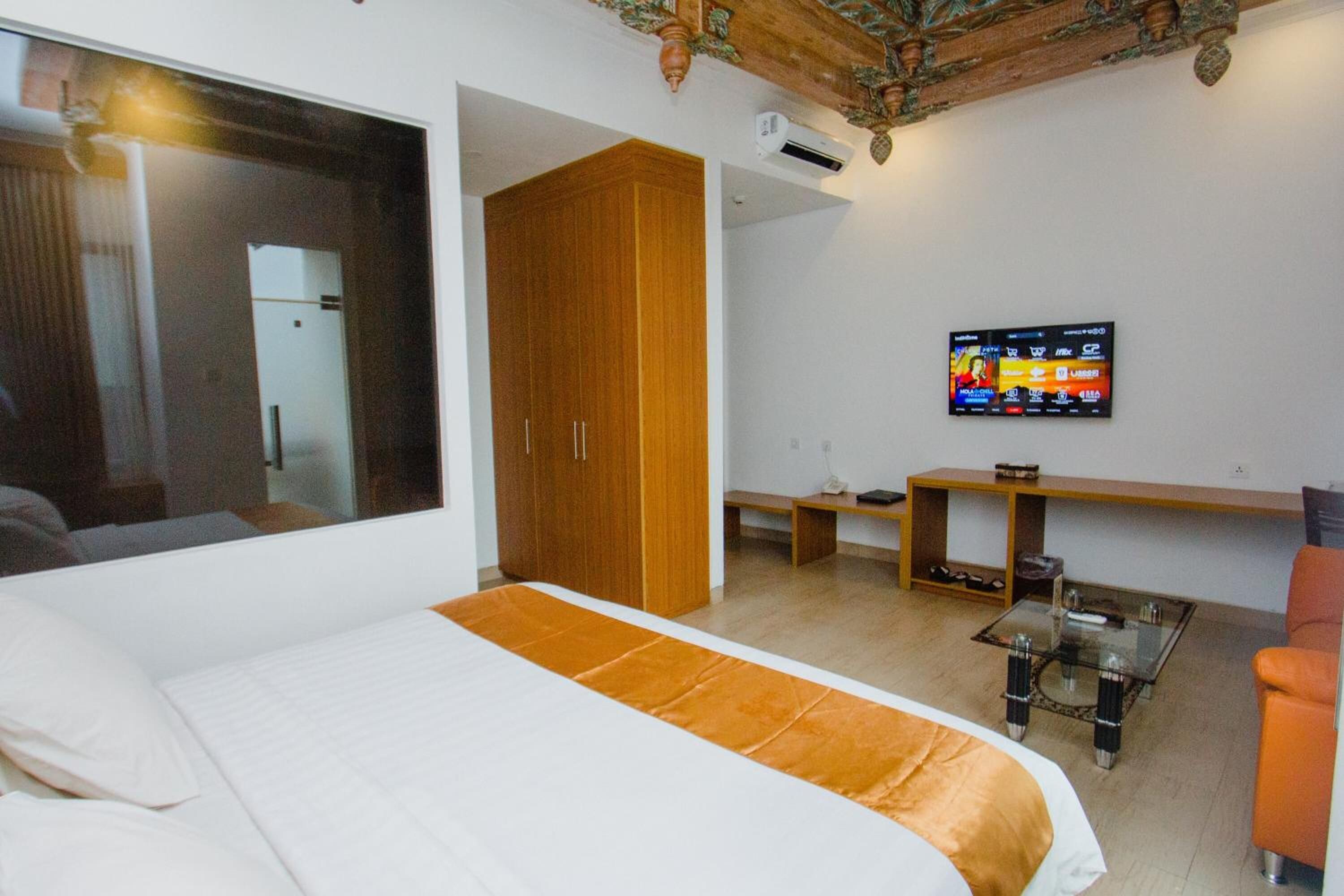 A bedroom with a large bed, a wooden wardrobe, a television on the wall, and a small table with a glass top.