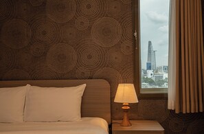 A hotel room with a bed, a nightstand with a lamp, a patterned wall, and a window with a view of a cityscape. - Mayfair Suites (Ho Chi Minh City)
