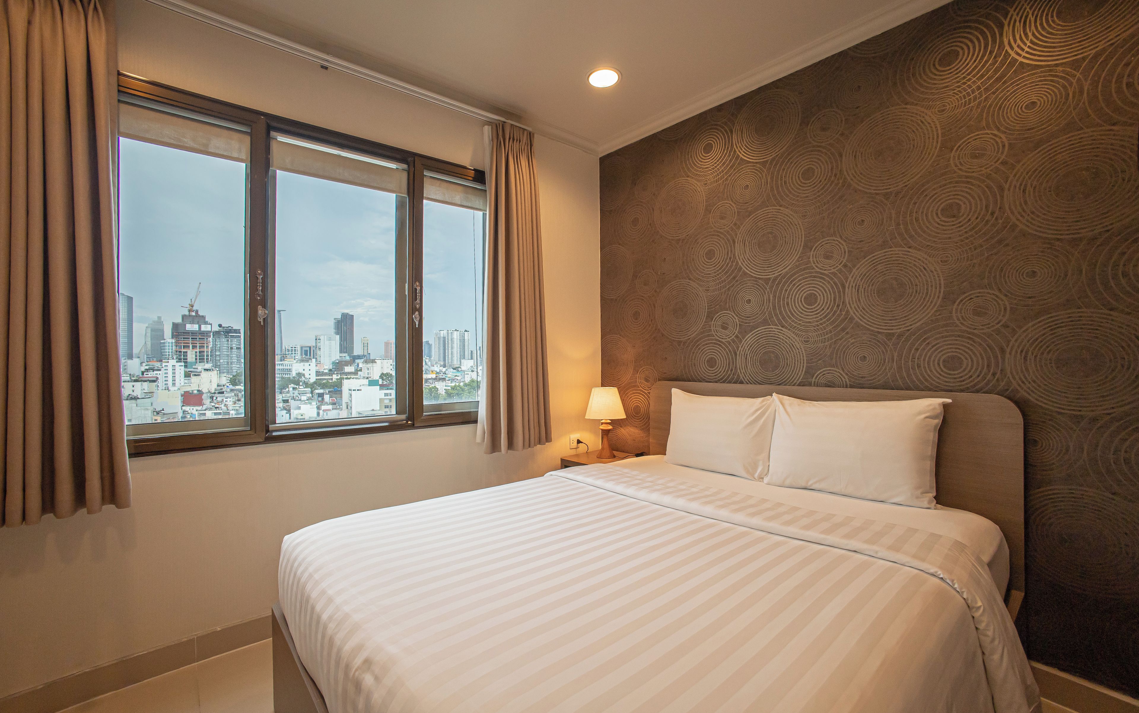 A hotel room with a bed, a nightstand, a lamp, a window with curtains, and a cityscape view.