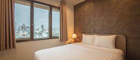 A hotel room with a bed, a nightstand, a lamp, a window with curtains, and a cityscape view.