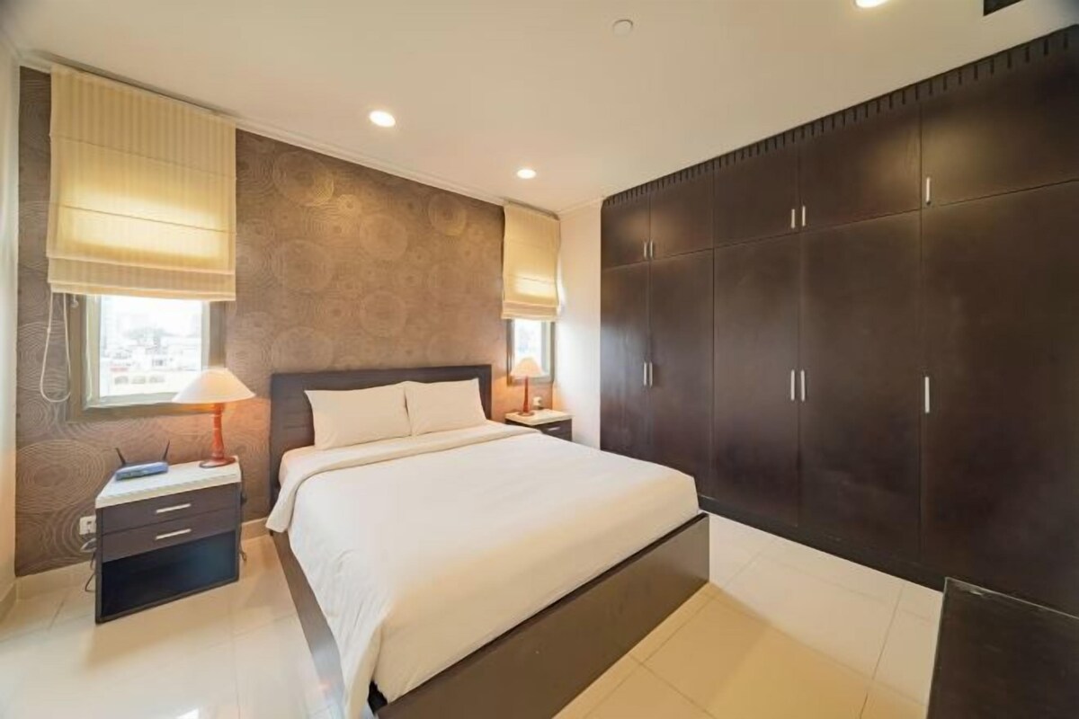 Quarto com cama grande, guarda-roupa de madeira escura, mesas de cabeceira e janela com persianas.