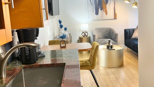 A modern kitchen with a marble countertop, a coffee maker, and a sink. There is a dining area with a yellow chair and a round table. A ceiling fan and a painting are visible on the wall. - Miami Beach HomeStay (North Miami)