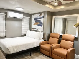 Un dormitorio con una cama, un sillón de cuero marrón, un equipo de aire acondicionado, un ventilador de techo y un cuadro abstracto grande.