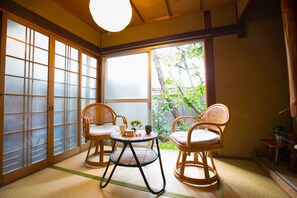 Une pièce de style japonais traditionnel, avec un sol recouvert de tatamis, une table en tatami, deux chaises en osier et une porte coulissante en verre donnant sur un jardin.