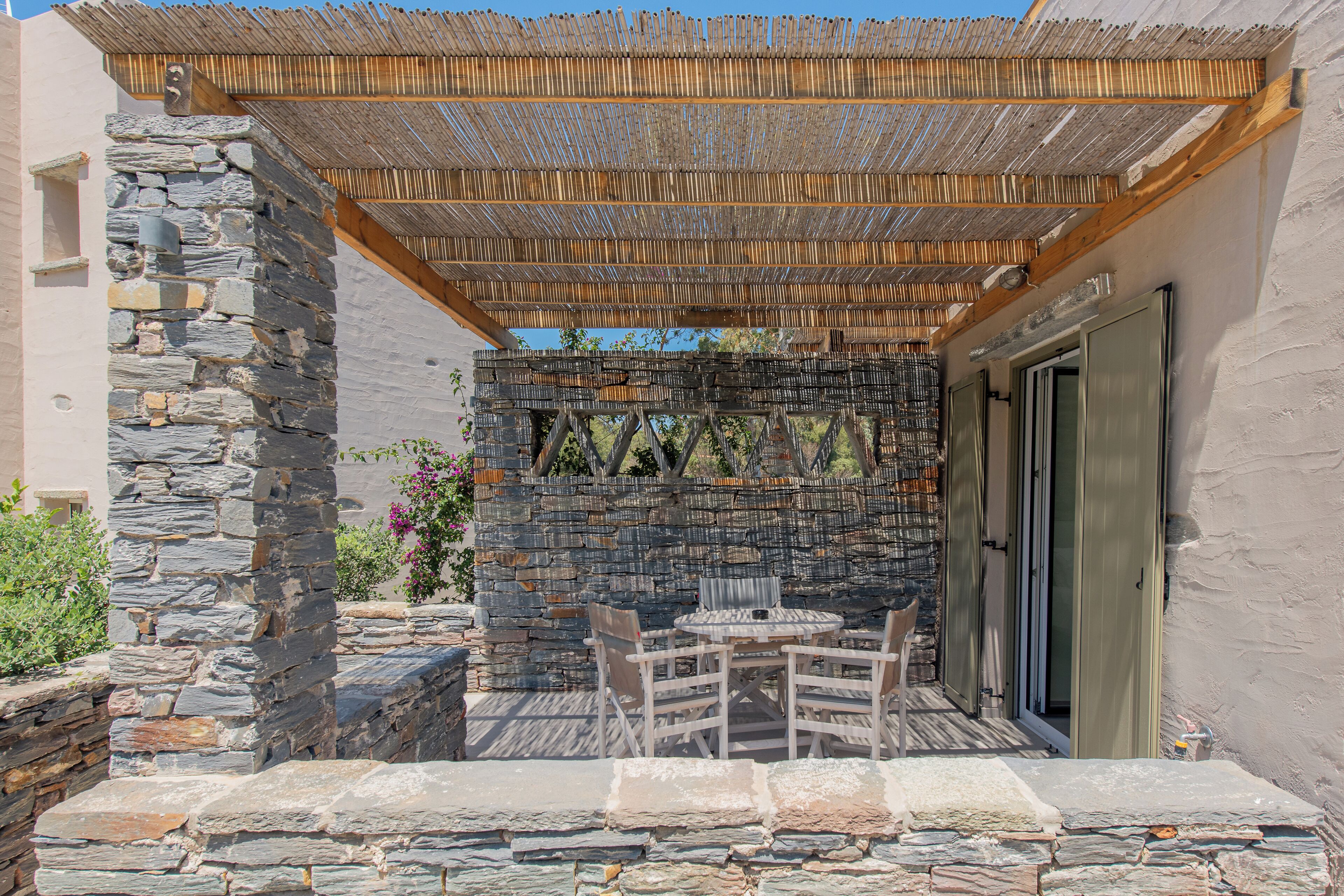 A stone wall with a wooden pergola, a table with chairs, and a door.