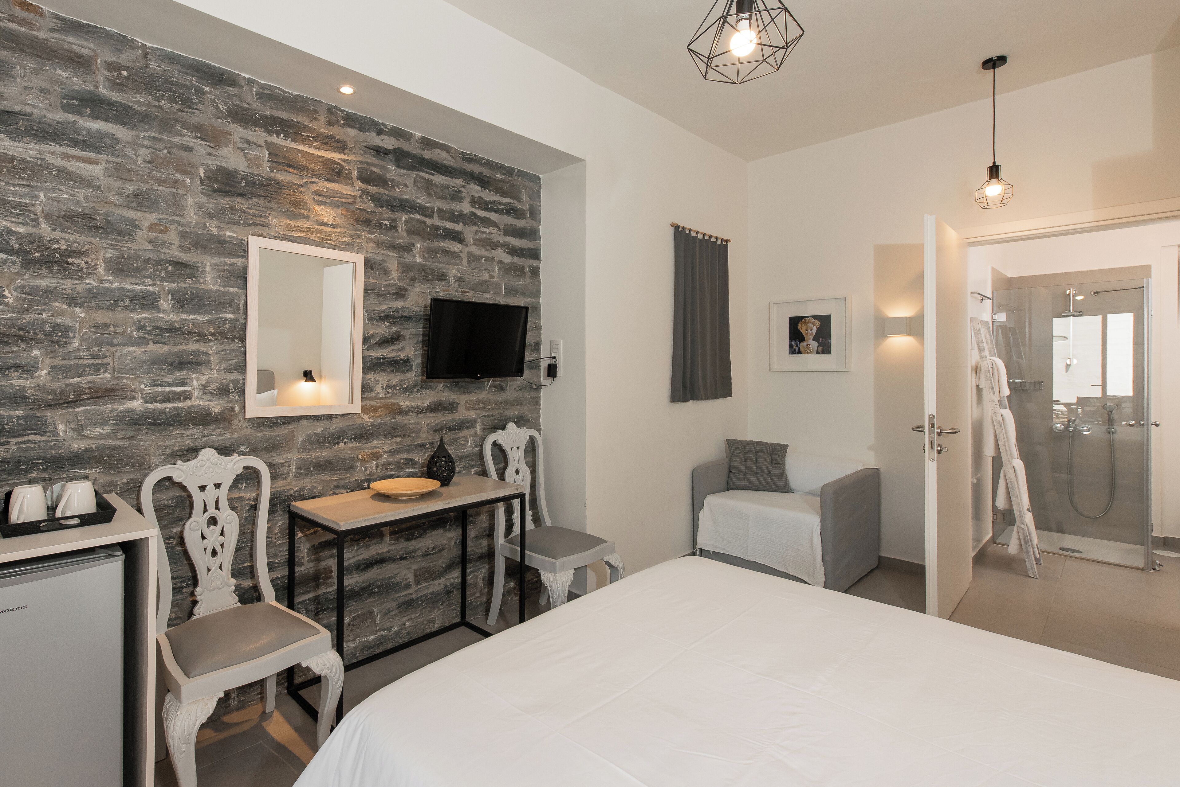 Une chambre d’hôtel moderne avec un mur en pierre, un lit, un bureau avec des chaises, une télévision et un espace douche.