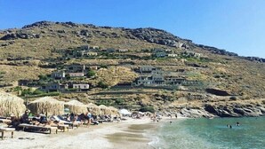 A beach with thatched umbrellas and sun loungers, a clear blue sea, and houses on a hillside in the background. - Vourkari Village (Kea)