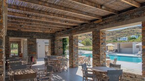 An outdoor patio area with stone columns, wooden tables, and chairs, overlooking a swimming pool and a hillside. - Vourkari Village (Kea)