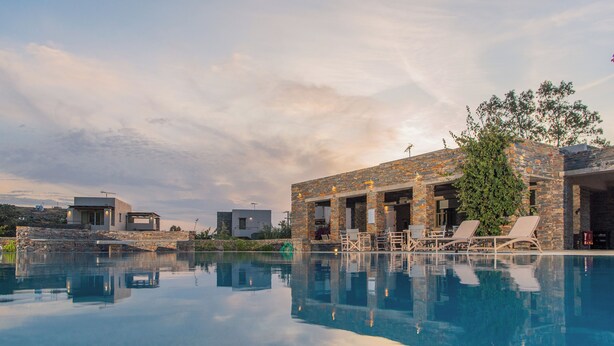 A modern pool area with a stone building, lounge chairs, and a clear pool reflecting the sky. - Vourkari Village (Kea)